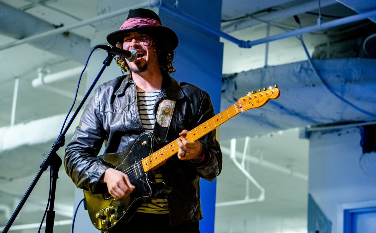Kenny Pirog Wearing A Leather Jacket And Fedora And Playing An Electric Guitar While Singing