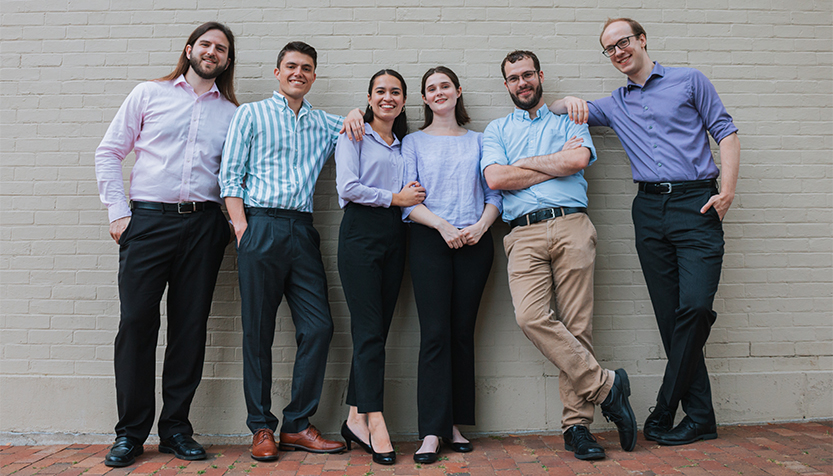 Ars Gratia Populi Group Members In Blue Shirts And Neutral Pants Against A White Brick Wall