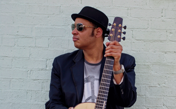 Raul Midon With His Guitar Kneeling In Front Of A White Wall