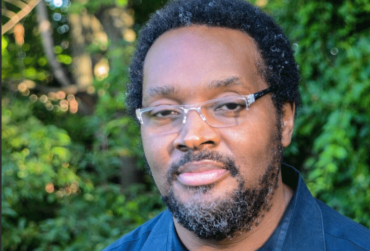 Keith Clark Wearing A Blue Shirt And Glasses In Front Of Greenery