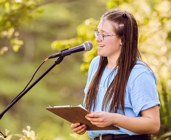 Emma Ash In A Light Blue Shirt Speaking At A Microphone