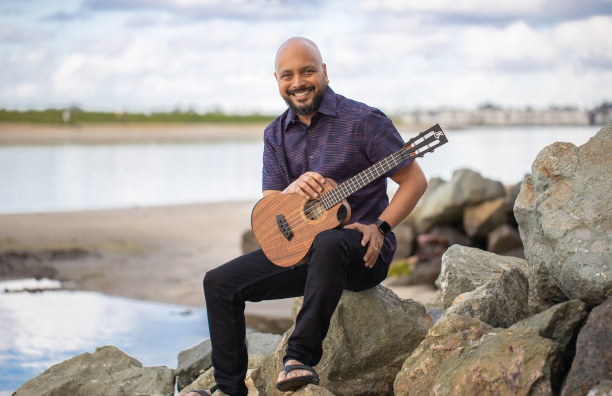 Steven Espaniola Sitting On Rocks By The Beach With His Ukulele